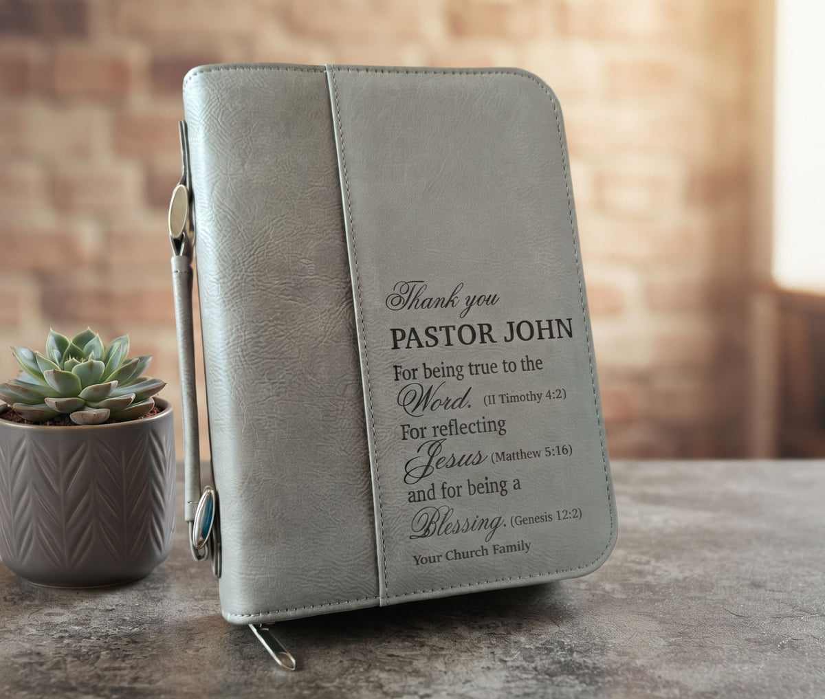 A personalized zippered Bible cover in textured light gray faux leather, featuring printed black text on the front pocket. It is styled on a stone tabletop next to a small potted succulent with a blurred brick wall background.