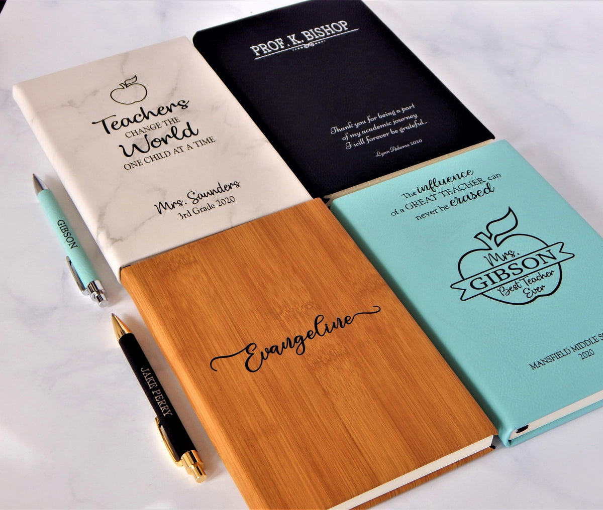 Four personalized stationery items arranged on a white marble surface: a white marble notebook with a teachers quote, a black notebook with a quote, a bamboo notebook with the name Evangeline, and a light blue hardcover notebook with a quote and apple logo. Two pens are also visible.