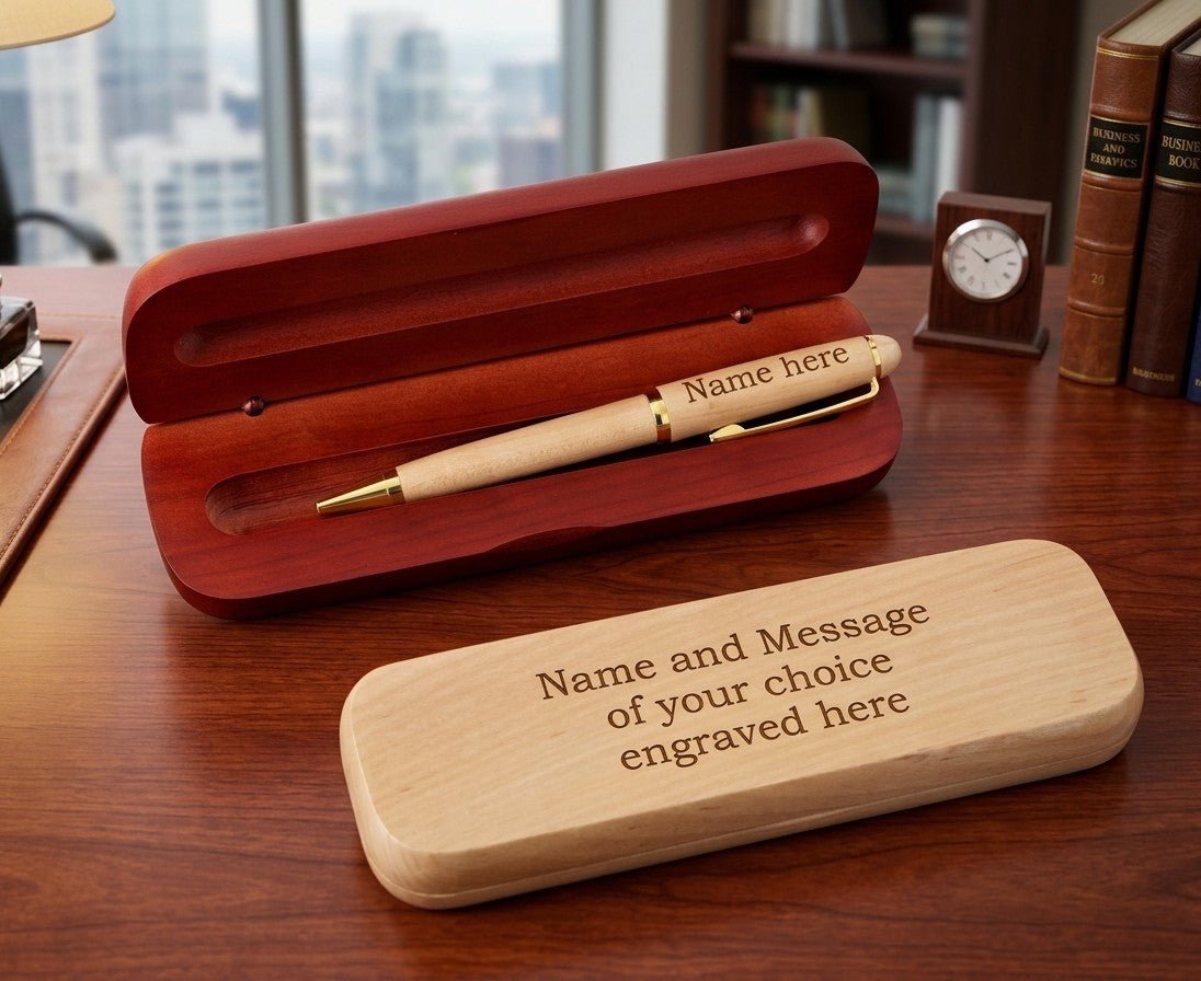 A personalized wooden pen gift set displayed on a wooden desk, featuring an open reddish-brown wooden presentation box with a light wood pen inside, and a matching engraved light wood lid in the foreground.