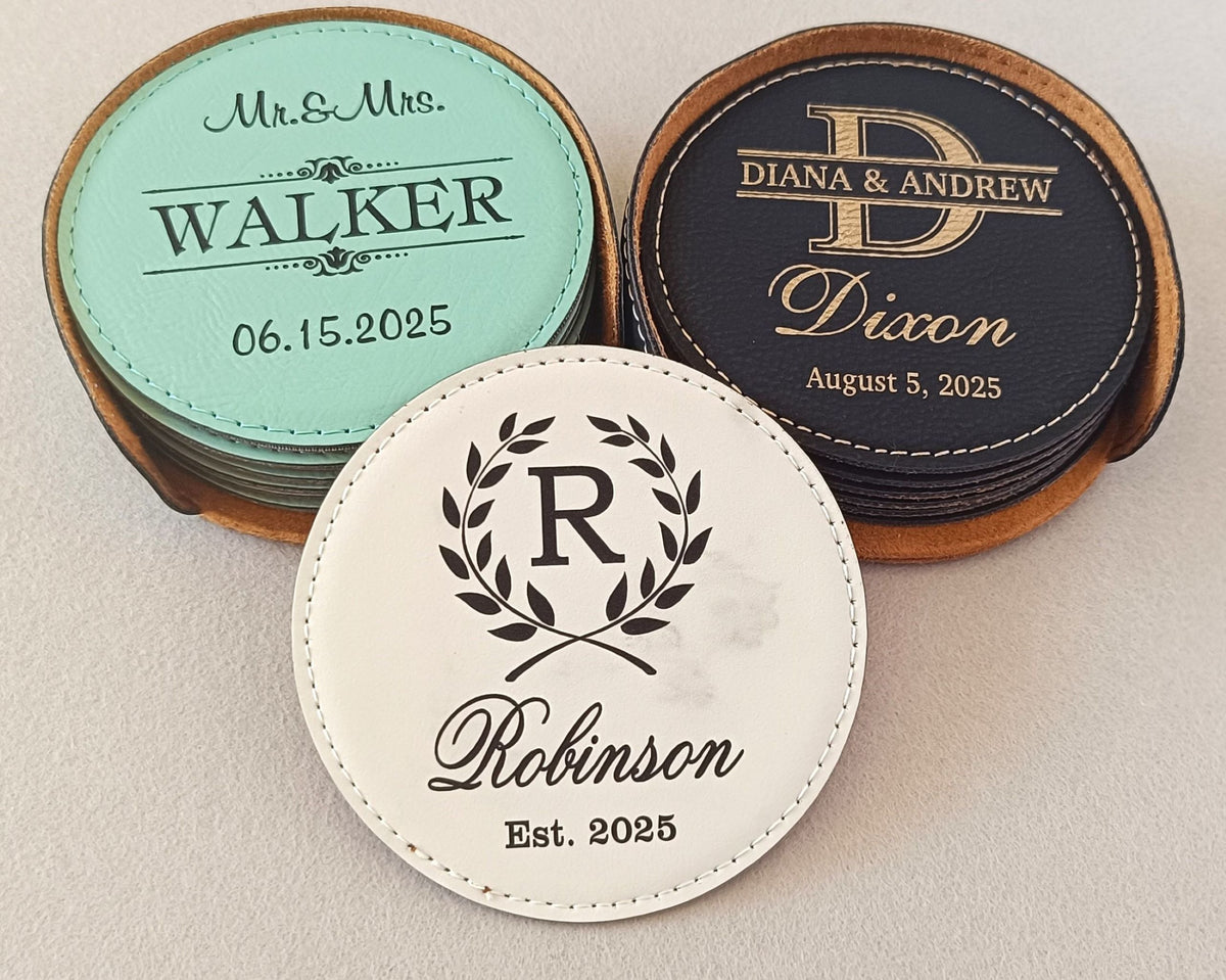 Three personalized round coasters in different colors and materials, displayed in their wooden holders on a light beige surface. The coasters feature elegant script and monogram designs with names and dates.